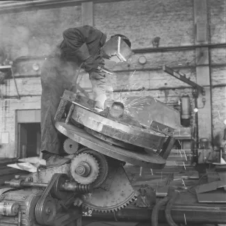 Mustavalkoinen kuva konepajan sisältä jossa työntekijä hitsaa monimutkaisen näköistä laitetta