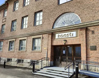 punatiilinen kolmikerroksinen rakennus jossa on paljon pieniruutuisia ikkunoita puolikaaren muotoisen ikkunan alla on kylrri jossa lukee konsti.